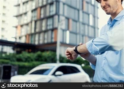 Businessman with smartwatch at modern charging station for electric vehicle with background of residential buildings as concept for progressive lifestyle of using eco-friendly as alternative energy.. Progressive businessman with smartwatch at public charging station for EV car.