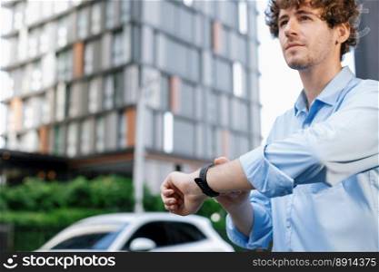 Businessman with smartwatch at modern charging station for electric vehicle with background of residential buildings as concept for progressive lifestyle of using eco-friendly as alternative energy.. Progressive businessman with smartwatch at public charging station for EV car.