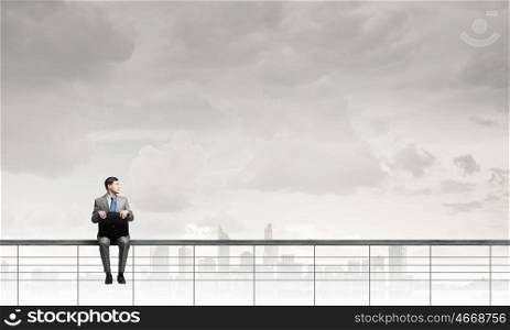 Businessman with suitcase. Young businessman sitting on parapet with briefcase in hands
