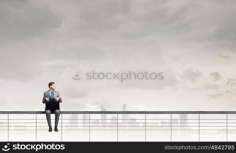 Businessman with suitcase. Young businessman sitting on parapet with briefcase in hands