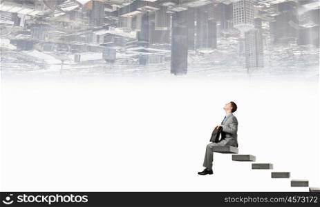 Businessman with suitcase. Young businessman sitting on stairs with briefcase in hands and city reflecting in sky