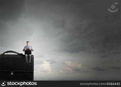 Businessman with suitcase. Young smiling businessman sitting on giant briefcase