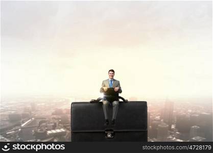Businessman with suitcase. Young smiling businessman sitting on giant briefcase
