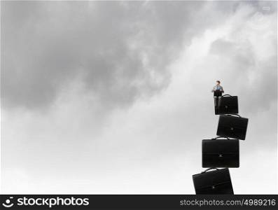 Businessman with suitcase. Young smiling businessman sitting on stack of briefcases