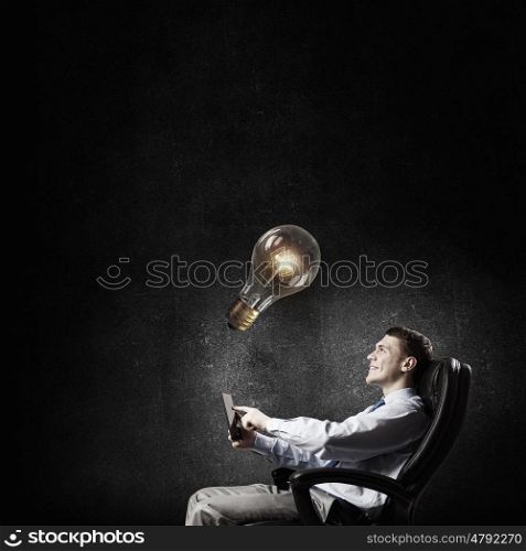 Businessman with tablet. Young businessman in armchair using tablet pc looking at glass lightbulb
