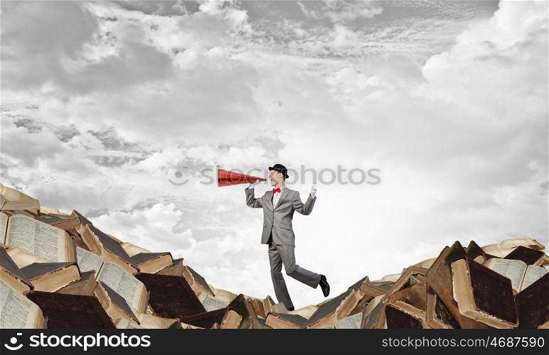 Businessman with trumpet. Young businessman on pile of books screaming emotionally in paper trumpet