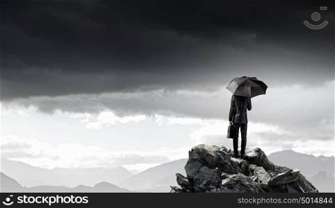 Businessman with umbrella. Back view of businessman with umbrella and suitcase standing on mountain top