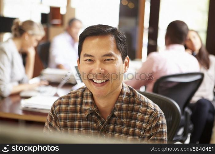 Businessman Working At Desk With Meeting In Background