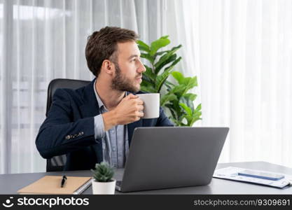 Businessman working on his laptop and drinking coffee at his desk in workspace. Smart executive taking coffee break after researching financial data and marketing plan on his corporate laptop. Entity. Businessman working on his laptop and drinking coffee at his desk. Entity