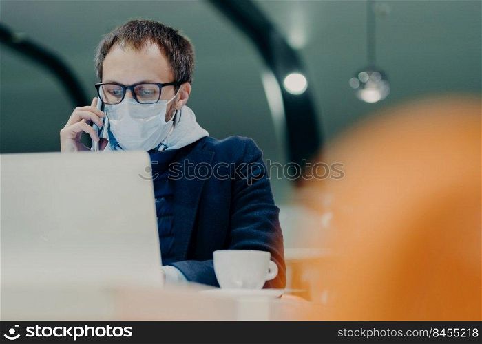 Businessman works remotely from cafeteria, concentrated at display of laptop computer, has telephone talk, wears medical mask on face as protection against flu or coronavirus epidemy. Smart working