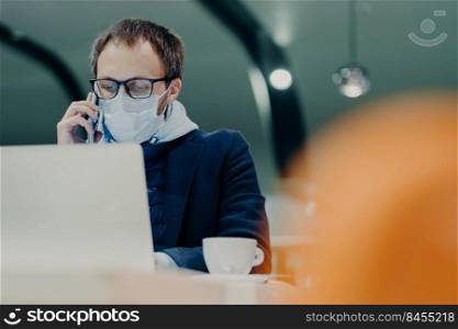 Businessman works remotely from cafeteria, concentrated at display of laptop computer, has telephone talk, wears medical mask on face as protection against flu or coronavirus epidemy. Smart working