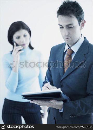 Businessman writing on a personal organizer with a businesswoman standing behind him holding a mobile phone