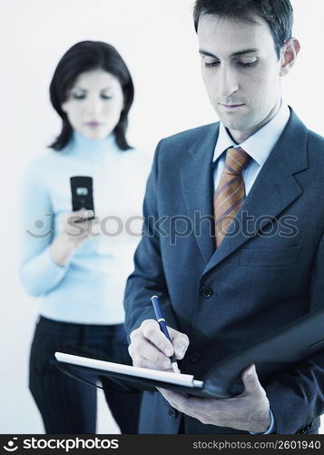 Businessman writing on a personal organizer with a businesswoman standing behind him holding a mobile phone