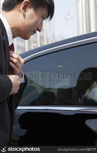 Businessmen straightening tie using reflection in car window.