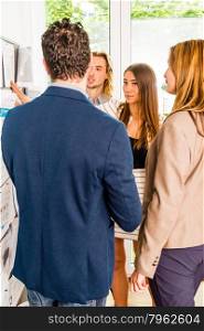 Businesspeople looking at bulletin board in office and discussing designs pinned at it. Mixed caucasian group rather casual, might be a startup comany or a creative agency.