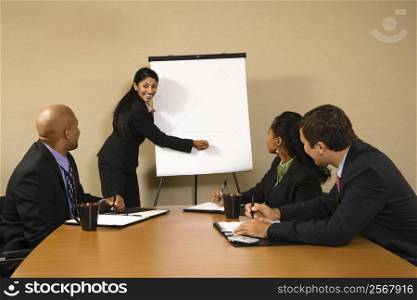 Businesspeople sitting at conference table smiling while businesswoman gives presentation.