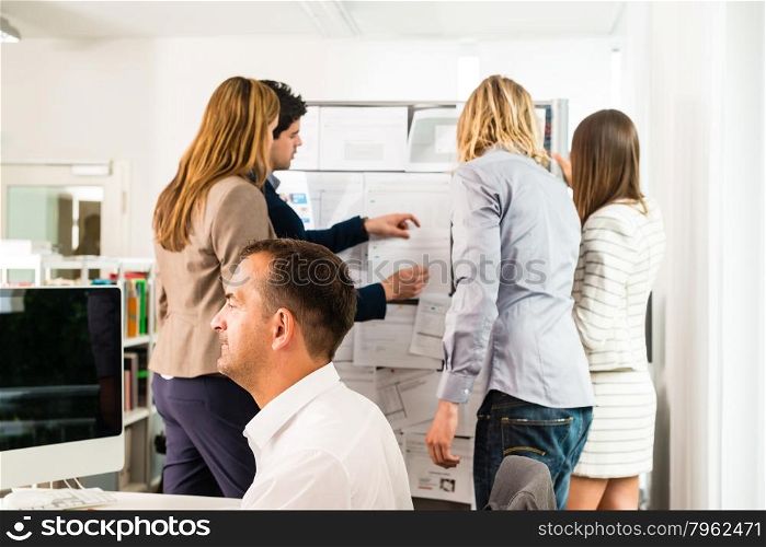 Businesspeople working in the office - some of them looking at bulletin board and discussing designs pinned at it, another guy in the front is working with several computer screens. Mixed caucasian group rather casual, might be a startup comany or a creative agency.