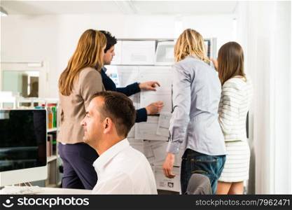Businesspeople working in the office - some of them looking at bulletin board and discussing designs pinned at it, another guy in the front is working with several computer screens. Mixed caucasian group rather casual, might be a startup comany or a creative agency.