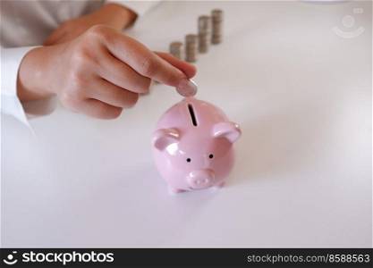 Businessperson Inserting Coins In Piggy Bank With Stack Of Coins Over The Desk 