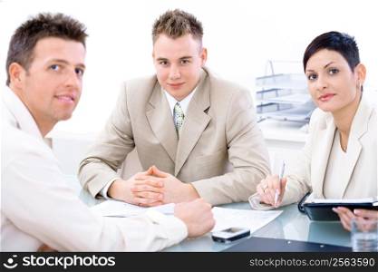 Businessteam of three working together, sitting around a desk, looking at camera.
