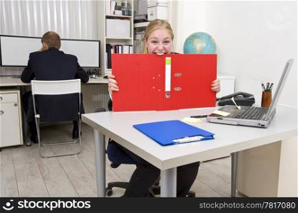 Businesswoman biting a dossier out of frustration, with an oblivious colleague in the background