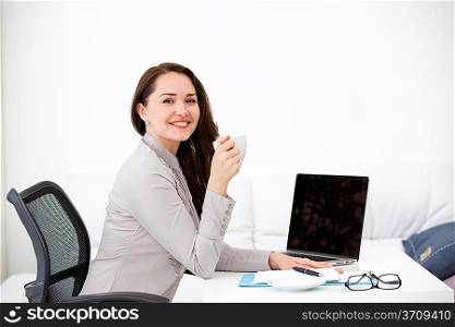 Businesswoman drinking coffee at desk, looking at camera, smiling in the office