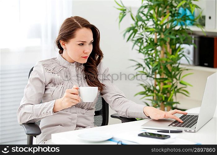 Businesswoman drinking coffee at desk, looking at laptop screen, smiling in the office