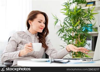 Businesswoman drinking coffee at desk, looking at laptop screen, smiling in the office