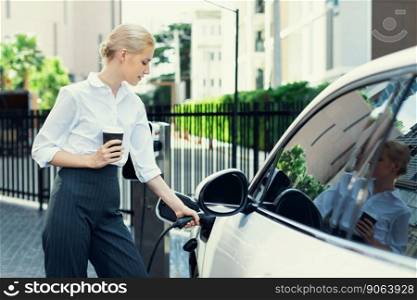 Businesswoman drinking coffee, leaning on electric vehicle recharging at public charging station with residential apartment condos building in background as progressive lifestyle by eco-friendly car.. Progressive woman with coffee while charging EV car with residential buildings.