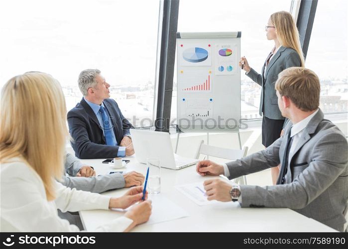 Businesswoman giving presentation using financial diagrams at flipchart to colleagues in office at meeting. Businesswoman giving presentation