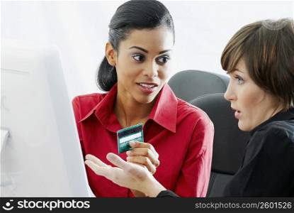 Businesswoman holding a credit card with another sitting beside her in front of a computer