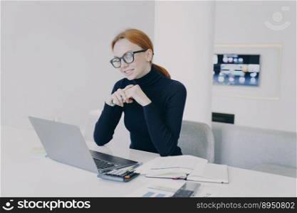 Businesswoman in glasses has online business video conference on quarantine and smiling at camera. White woman at workplace in office. Positive attractive girl entrepreneur is working at laptop.. Businesswoman in glasses has online business video conference and smiling at camera.