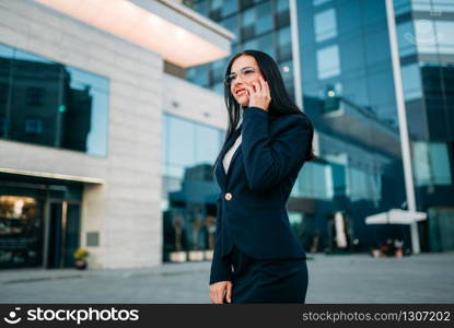 Businesswoman in suit talks by mobile phone outdoor, business center on background. Modern financial building, cityscape. Successful female businessperson