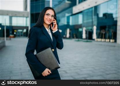 Businesswoman in suit talks by mobile phone outdoor, business center on background. Modern financial building, cityscape. Successful female businessperson. Businesswoman in suit talks by phone outdoor