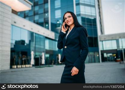Businesswoman in suit talks by mobile phone outdoor, business center on background. Modern financial building. Successful female businessperson. Businesswoman in suit talks by phone outdoor