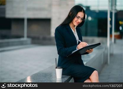 Businesswoman in suit writing in notebook outdoor, business center on background. Modern financial building, cityscape. Successful female businessperson