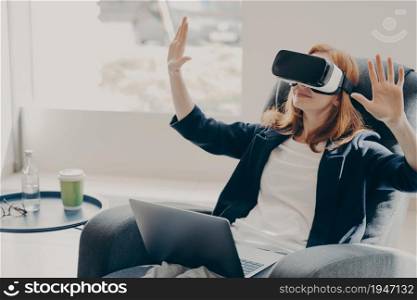 Businesswoman in virtual reality glasses using VR goggles, testing innovative method for business, sitting on armchair with opened laptop, interacting with digital interface. Cyberspace experience. Businesswoman in virtual reality glasses testing innovative method for business, working remotely
