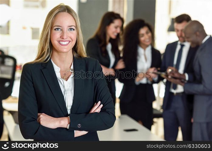Businesswoman leader looking at camera in modern office with multi-ethnic businesspeople working at the background. Teamwork concept. Blonde caucasian woman