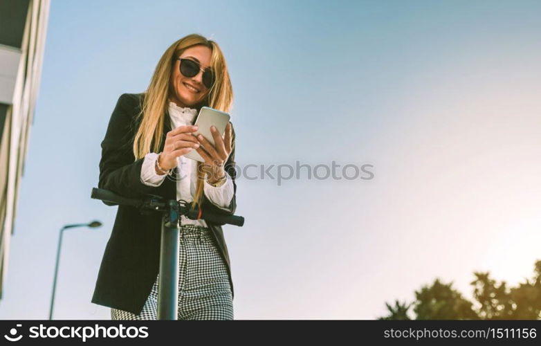 Businesswoman leaning on her electric scooter looking at the mobile in the street