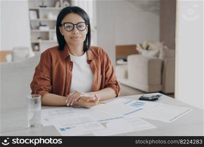 Businesswoman looking at camera, working on project at office desk with documents. Modern pretty female manager wearing glasses and casual clothes sitting on workplace, posing for business portrait.. Businesswoman wearing glasses looking at camera, working on project at office desk with documents