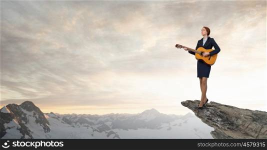 Businesswoman playing acoustic guitar. Cheerful businesswoman standing on rock edge and playing acoustic guitar