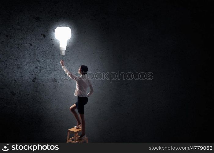 Businesswoman reaching light bulb. Back view of businesswoman standing on chair and reaching light bulb