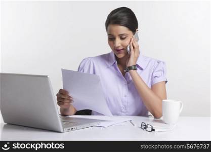 Businesswoman reading document while answering smart phone at desk against white background