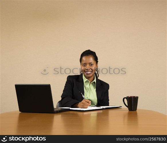 Businesswoman sitting at conference table with laptop computer and coffee cup smiling and writing in book.