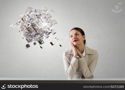 Businesswoman sitting at desk. Happy attractive businesswoman sitting at table in office and 3D rendering cube figure