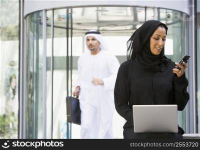 Businesswoman sitting outdoors by building with laptop holding cellular phone with businessman running in background (selective focus)