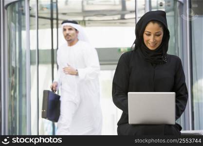 Businesswoman sitting outdoors by building with laptop smiling with businessman running in background (selective focus)