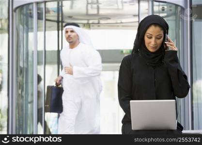 Businesswoman sitting outdoors by building with laptop using cellular phone with businessman running in background (selective focus)