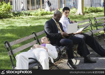 Businesswoman sleeping on a bench with two businessmen sitting beside her and looking at a laptop