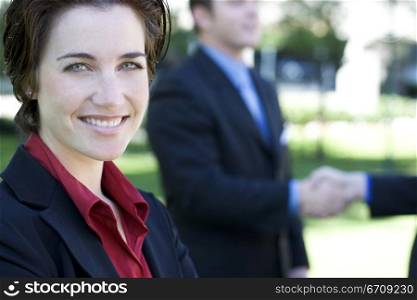 Businesswoman smiles and looks ahead while two businessmen shake hands